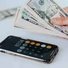 Dłoń trzyma banknoty dolarów nad smartfonem z otwartą aplikacją kalkulatora.